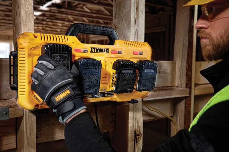 Een persoon met werkhandschoenen plaatst of verwijdert accu’s uit een gele DEWALT multi-accu oplader die aan een houten balk op een bouwplaats is bevestigd.