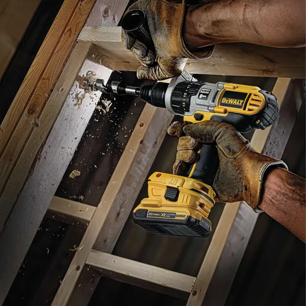 Een persoon met bruine werkhandschoenen gebruikt een draadloze DEWALT boormachine om een gat te boren in een houten balk, waarbij houtspaanders door het boren wegvliegen.