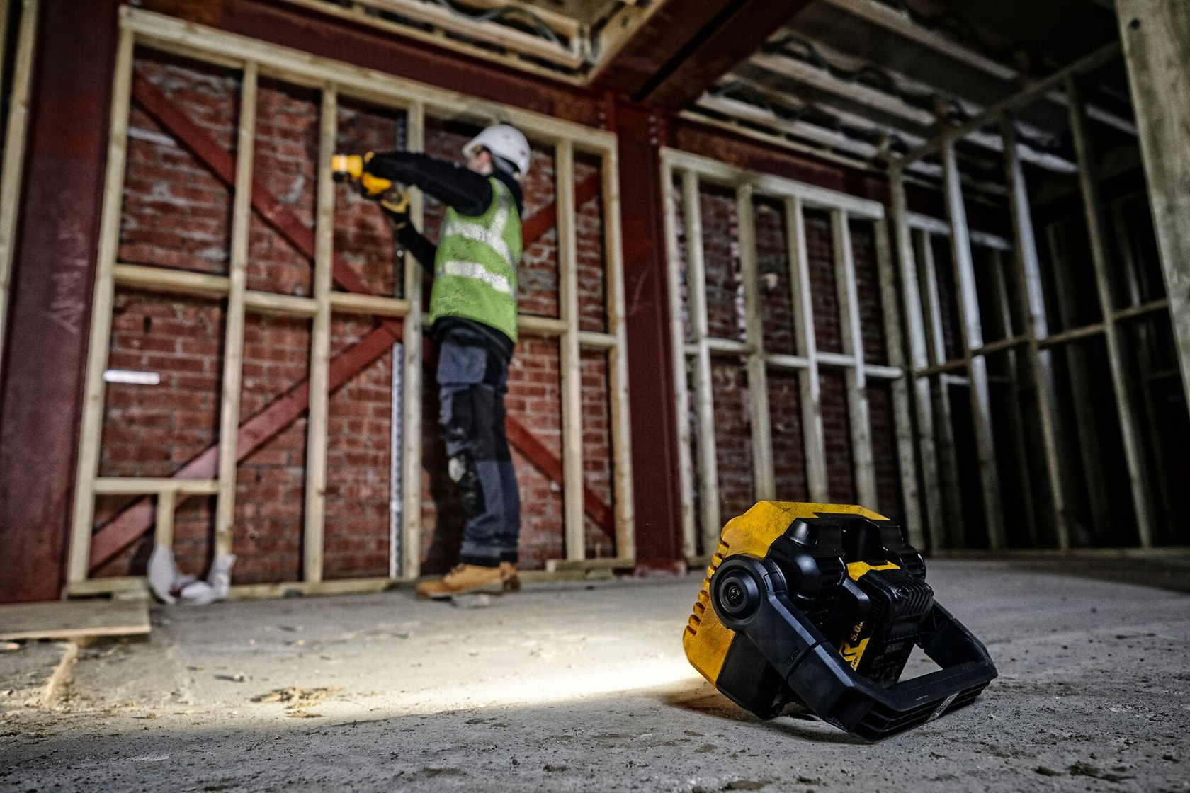 DEWALT DCL077 werklamp op de vloer van bouwplaats, werknemer op achtergrond bij houten frame.